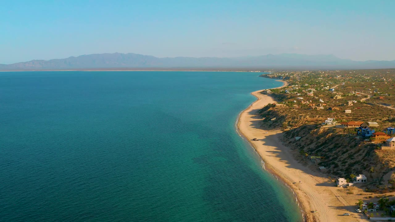 Wide aerial coastline with blue-green water, sand, and low coastal buildings.