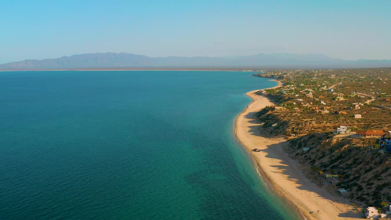 Aerial frame over El Sargento La Ventana, Mexico, with turquoise water on the left, a pale beach curving through the center, coastal homes on the right, distant mountains, and no visible people.