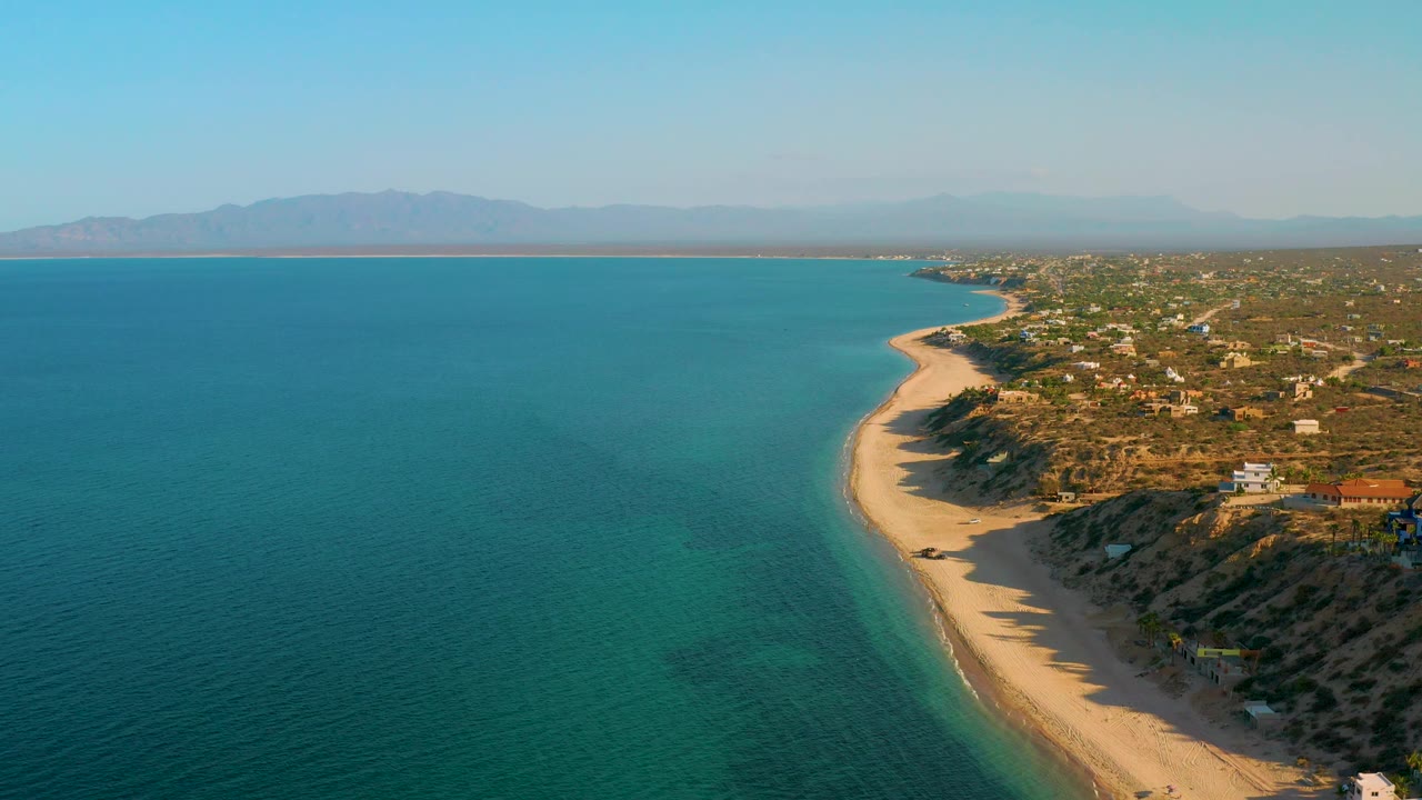 Drone-like aerial frame over El Sargento La Ventana, Mexico, looking along clear water and a sandy beach, with low homes and dry hills to the right and no visible people.