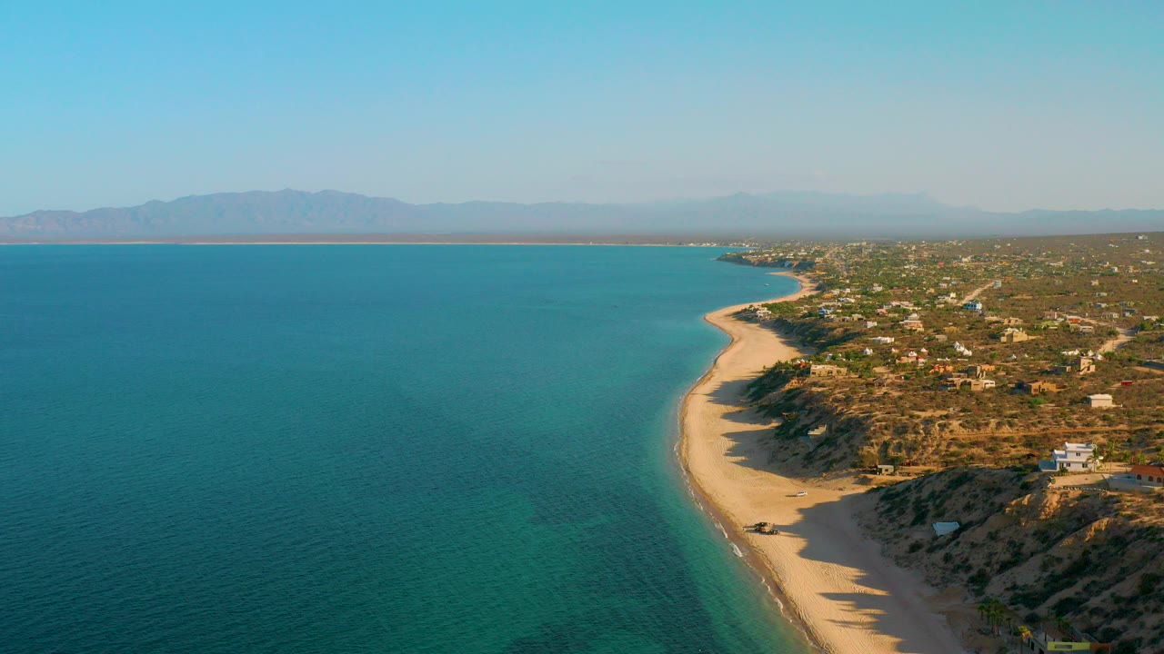 Opening aerial frame over El Sargento La Ventana, Mexico, with turquoise sea filling the left side, a pale beach curving along scrubby bluffs, scattered low homes to the right, distant mountains, and no visible people.