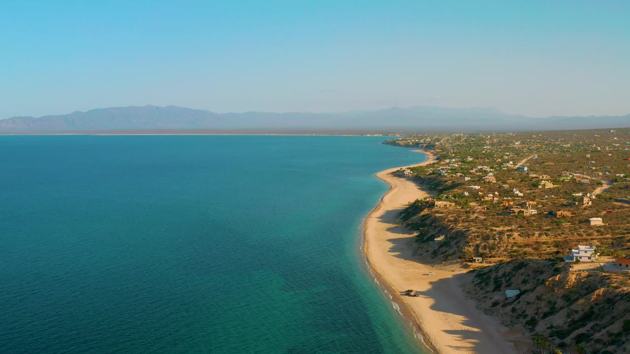 Two seconds in, the drone view shows more open turquoise water on the left, a bright sand strip bending through the center, scrubby coastal bluffs and scattered buildings on the right, a hazy mountain ridge beyond the gulf, and no visible people.