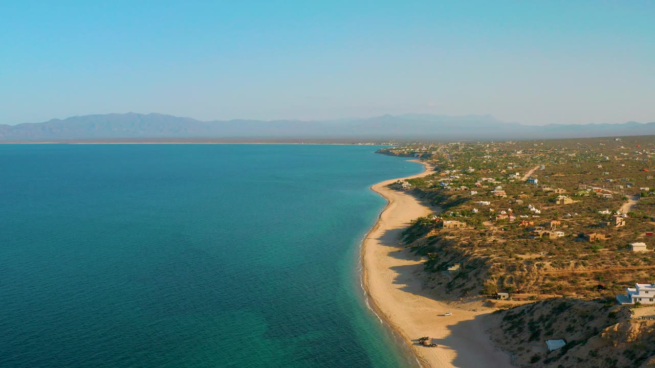 High aerial view over El Sargento La Ventana, Mexico, with blue water filling the left side, a curved sand beach below, scrubland and scattered buildings to the right, distant mountains, and no visible people.