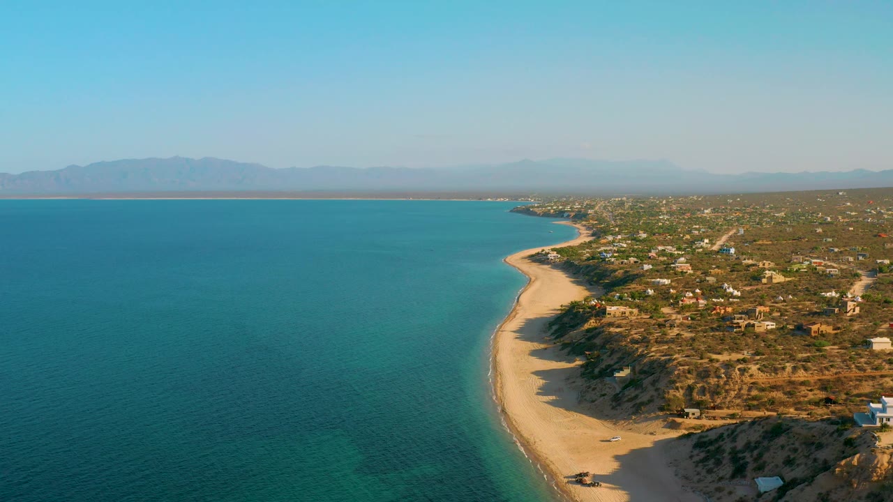 High view over El Sargento La Ventana, Mexico, with open blue water occupying most of the left frame, tan beach tracing the coast, dry land and scattered houses on the right, mountains in the distance, and no visible people.