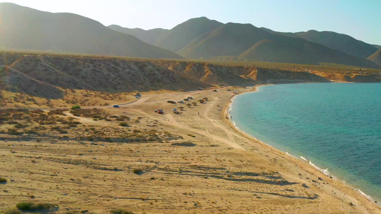 Aerial view at El Sargento La Ventana, Mexico, showing the shoreline slightly farther along, small parked vehicles near the sand, turquoise water at right, and no visible people.