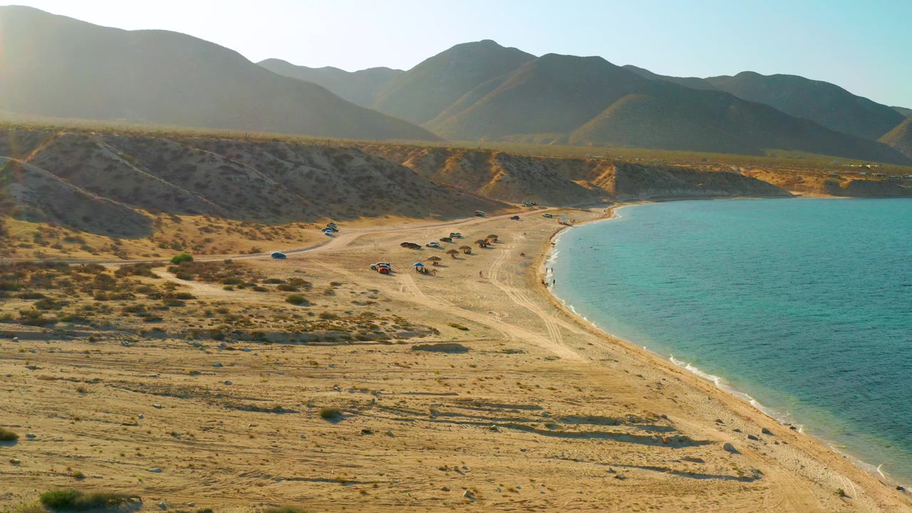 Mid-clip aerial view of El Sargento La Ventana, Mexico, with low sunlight washing the left side, a sandy beach below, parked vehicles near the shore, turquoise water, and no visible people.