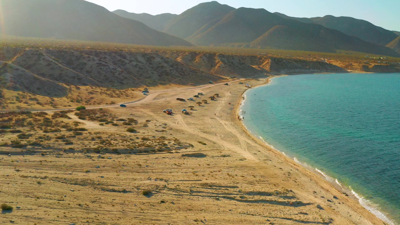 Aerial frame over El Sargento La Ventana showing dry sand, scrub, beach tracks, small parked vehicles by the curved shoreline, turquoise water, distant mountains, and no visible people.