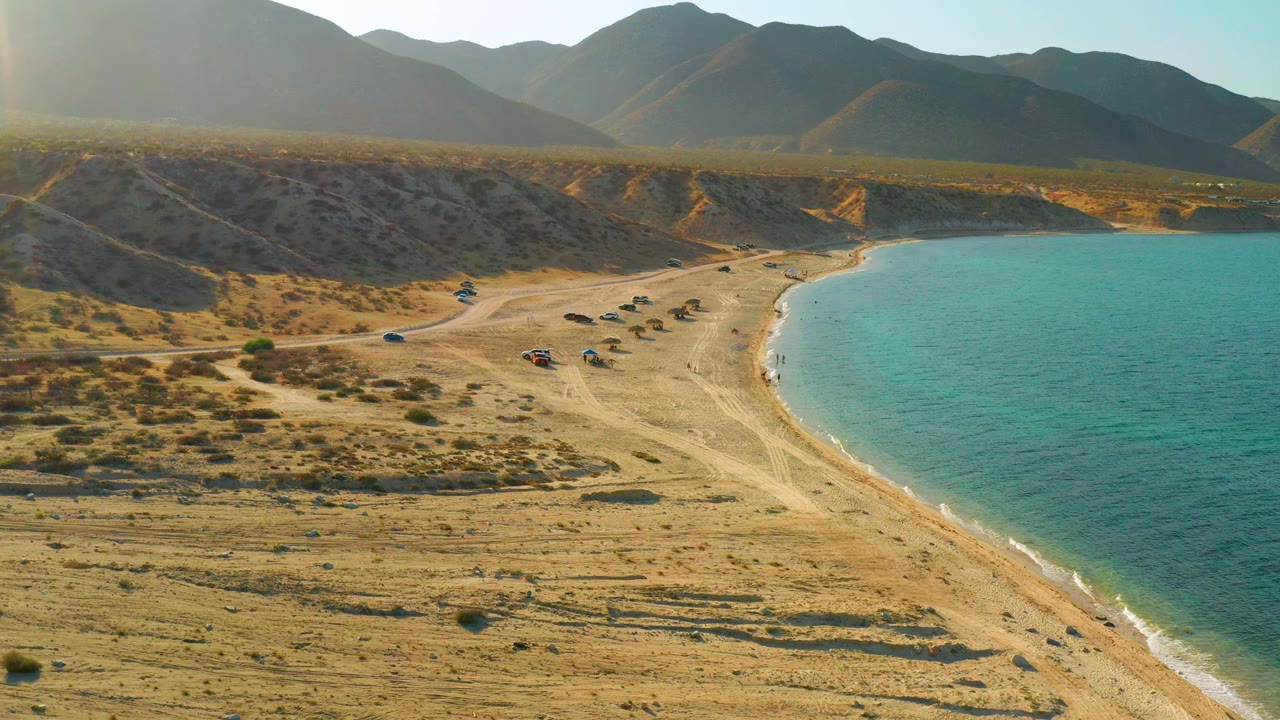 High drone view in El Sargento La Ventana with scrub-covered sand in front, a pale beach road, clustered parked vehicles near the waterline, blue-green ocean, mountains, and no visible people.