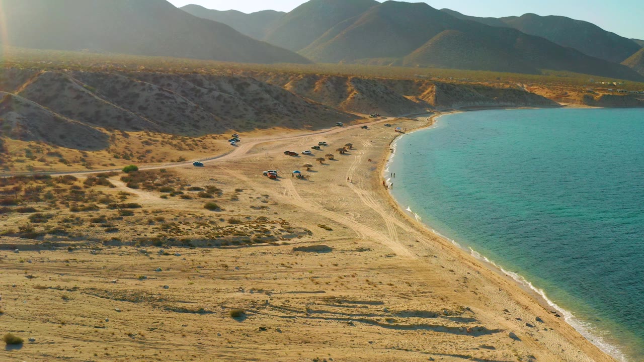 Aerial frame from El Sargento La Ventana with the turquoise sea occupying the right side, a narrow pale beach curve, scattered parked vehicles, dry ridges behind the shore, mountains, and no visible people.