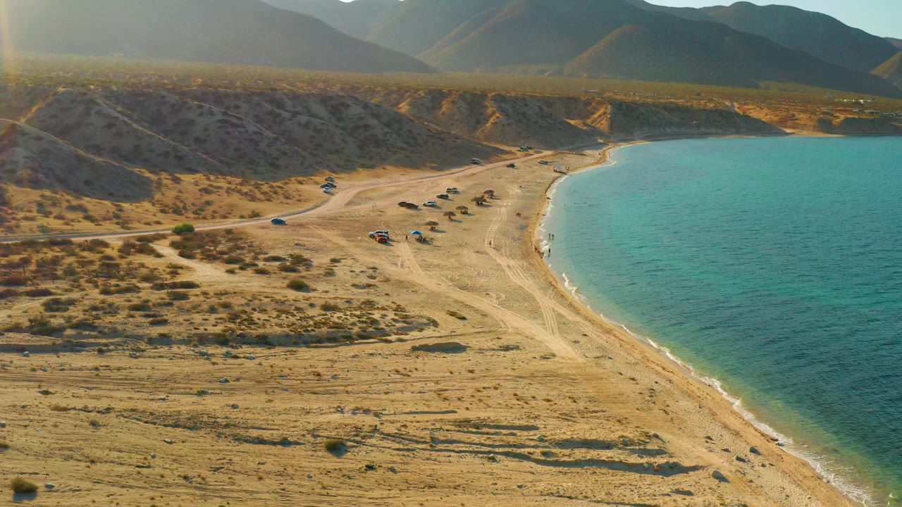 Mid-clip aerial view of El Sargento La Ventana with dirt access tracks crossing sandy scrub, small vehicles parked near the beach, calm turquoise water on the right, hazy mountains, and no visible people.