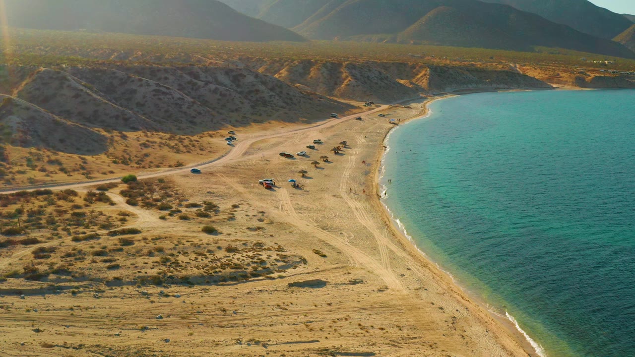 At 0:01, the drone-height view in El Sargento La Ventana, Mexico holds on the blue-green shoreline, pale sand, inland access road, scattered shrubs, small vehicles, mountains, and no visible people.