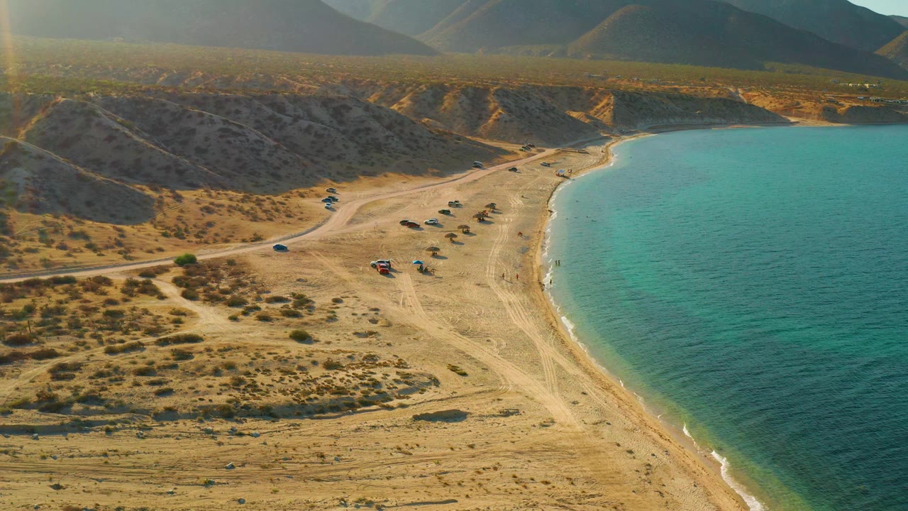 At 0:02, the frame shows El Sargento La Ventana's curved sandy edge with calm turquoise water to the right, dry brush and tracks inland, low hills beyond, parked vehicles, and no visible people.