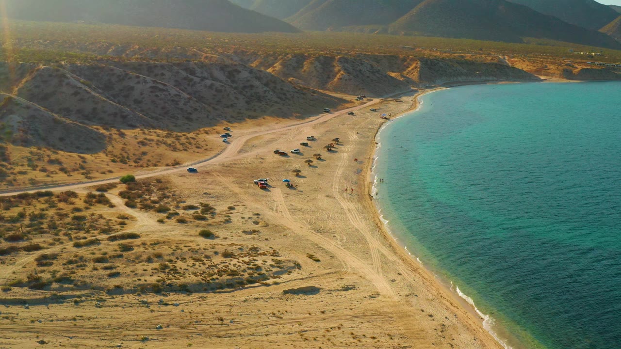 At 0:03, the aerial frame in El Sargento La Ventana, Mexico places shallow clear water along the right edge, tan beach through the center, vehicles near the shore, scrubland left, and no visible people.