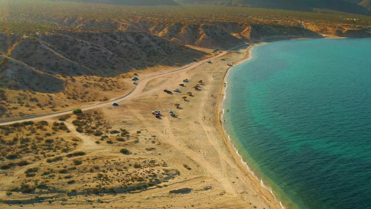 Aerial frame of El Sargento La Ventana coastline with the sea filling the right side, sandy beach and road through the center, dry hills behind, small beach objects, and no visible people.