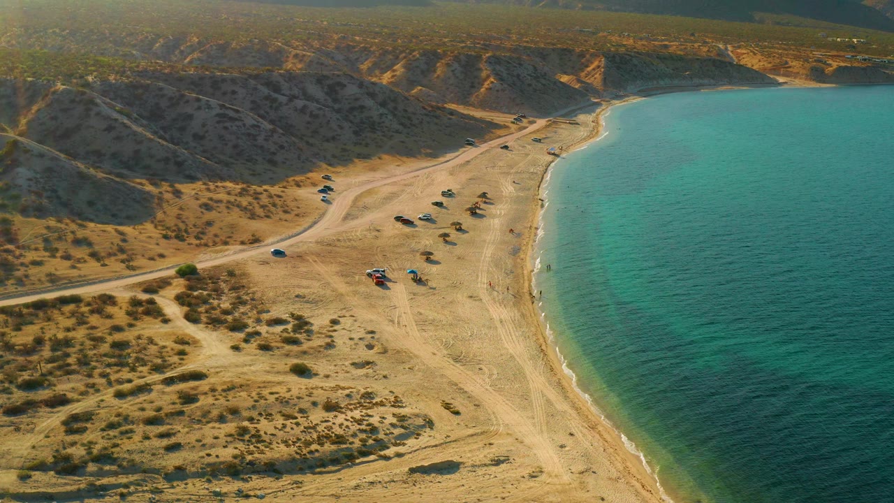 High oblique view of El Sargento La Ventana, Mexico, with clear turquoise water along a curved beach, arid hills above the shore, a winding access road, clustered small vehicles or shelters, and no visible people.
