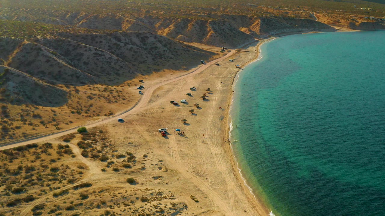 Oblique aerial view in El Sargento La Ventana, Mexico, with the shoreline curving past scattered vehicles or shade setups and no visible people.