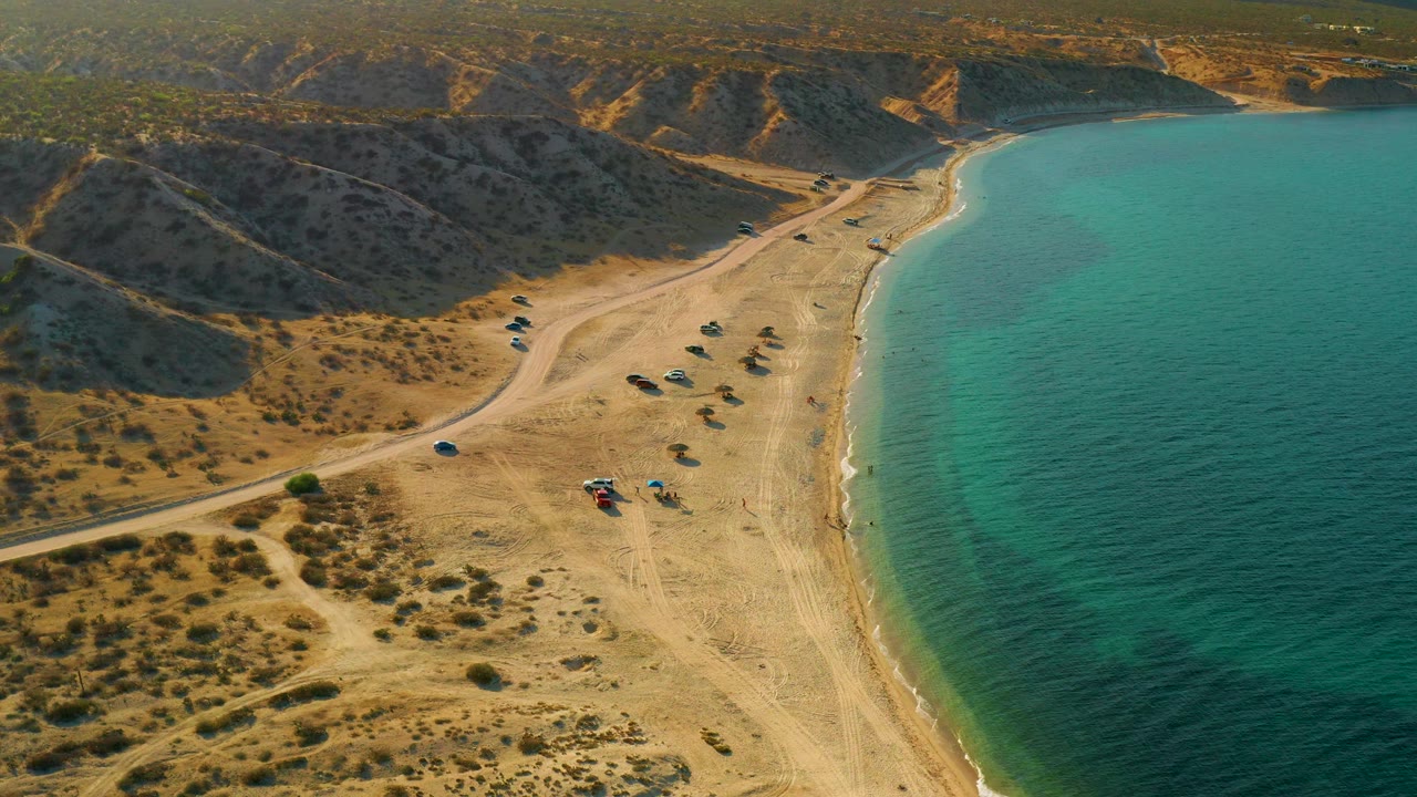 High aerial frame over El Sargento La Ventana, Mexico, with clear green water, a pale sand strip, a dirt road, brown hills, and no visible people.