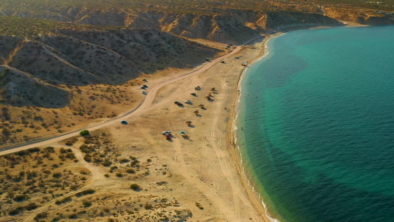 Aerial view in El Sargento La Ventana, Mexico, showing turquoise water along the right edge, sandy beach through the center, scrub foreground, and no visible people.