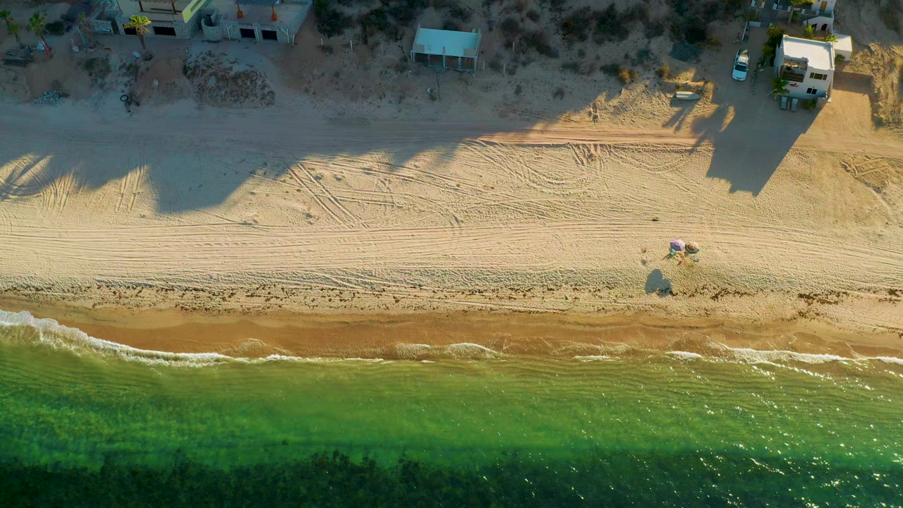Overhead view of El Sargento La Ventana shoreline with green water at the bottom, thin breaking waves, raked pale sand, beach structures at the top, and no visible people.