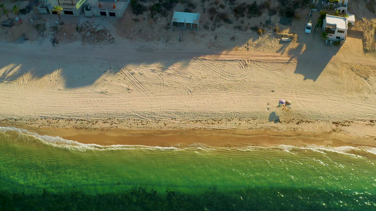 Top-down El Sargento La Ventana beach frame with sunlit sand, narrow foam along the waterline, green water in the lower half, shoreline buildings above, and no visible people.