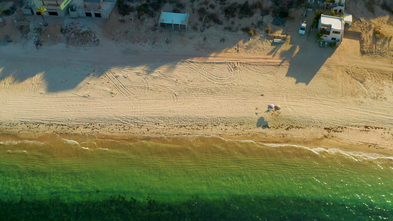 Aerial view over El Sargento La Ventana, Mexico, with green shallow water below, a sandy beach with tire tracks, small buildings near the top edge, and beachgoers visible by colorful shade.