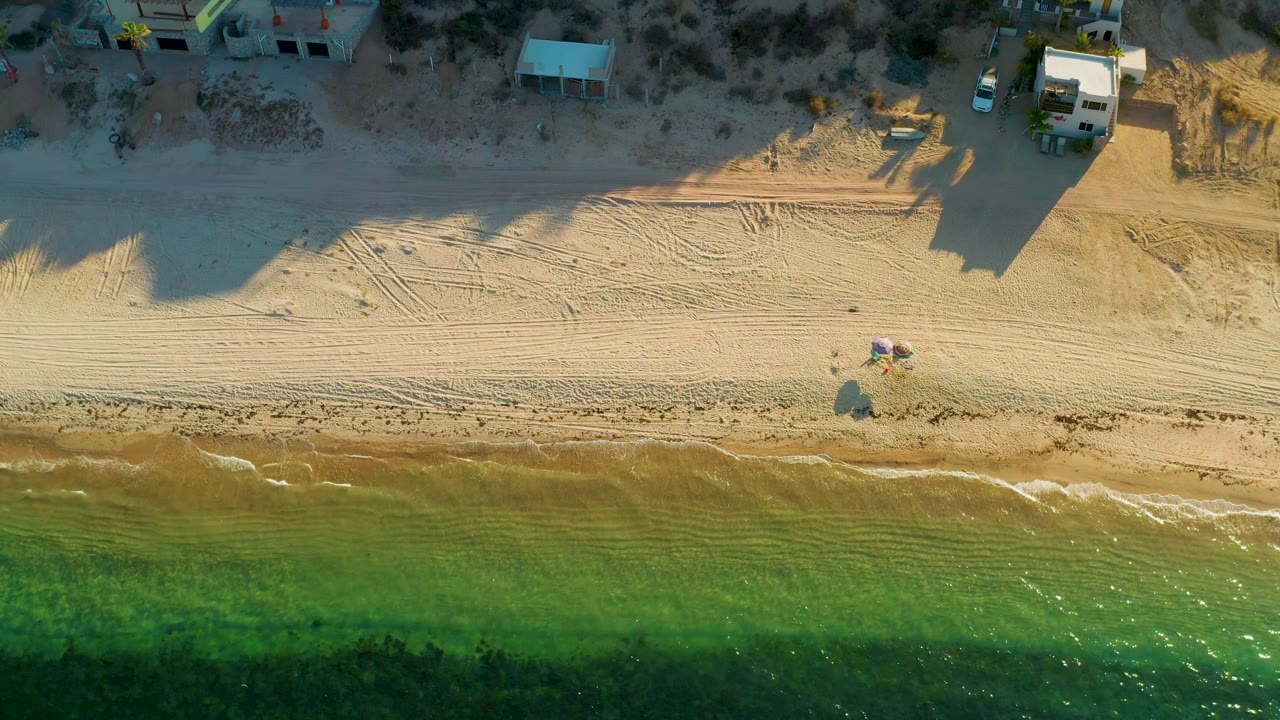 Drone view of sandy shoreline, small waves, beachgoers, and buildings.