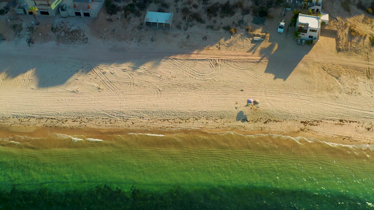 Overhead view in El Sargento La Ventana, Mexico, with a narrow surf line, broad pale sand, colorful beach shade near the lower sand area, and small structures along the upper beach edge.
