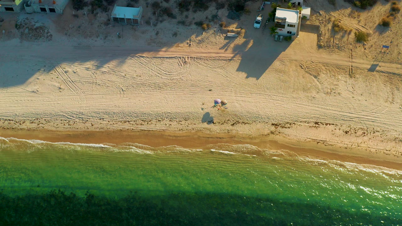 Top-down aerial frame over El Sargento La Ventana, Mexico, with green-blue water below, tire-marked sand in the center, small houses near the top, and no visible people.