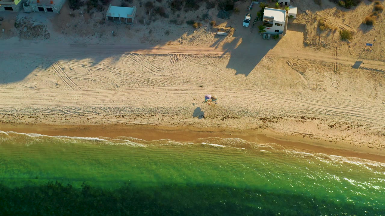 Aerial view in El Sargento La Ventana, Mexico, showing shallow green surf, a narrow brown wet-sand strip, patterned beach sand, house shadows, and no visible people.