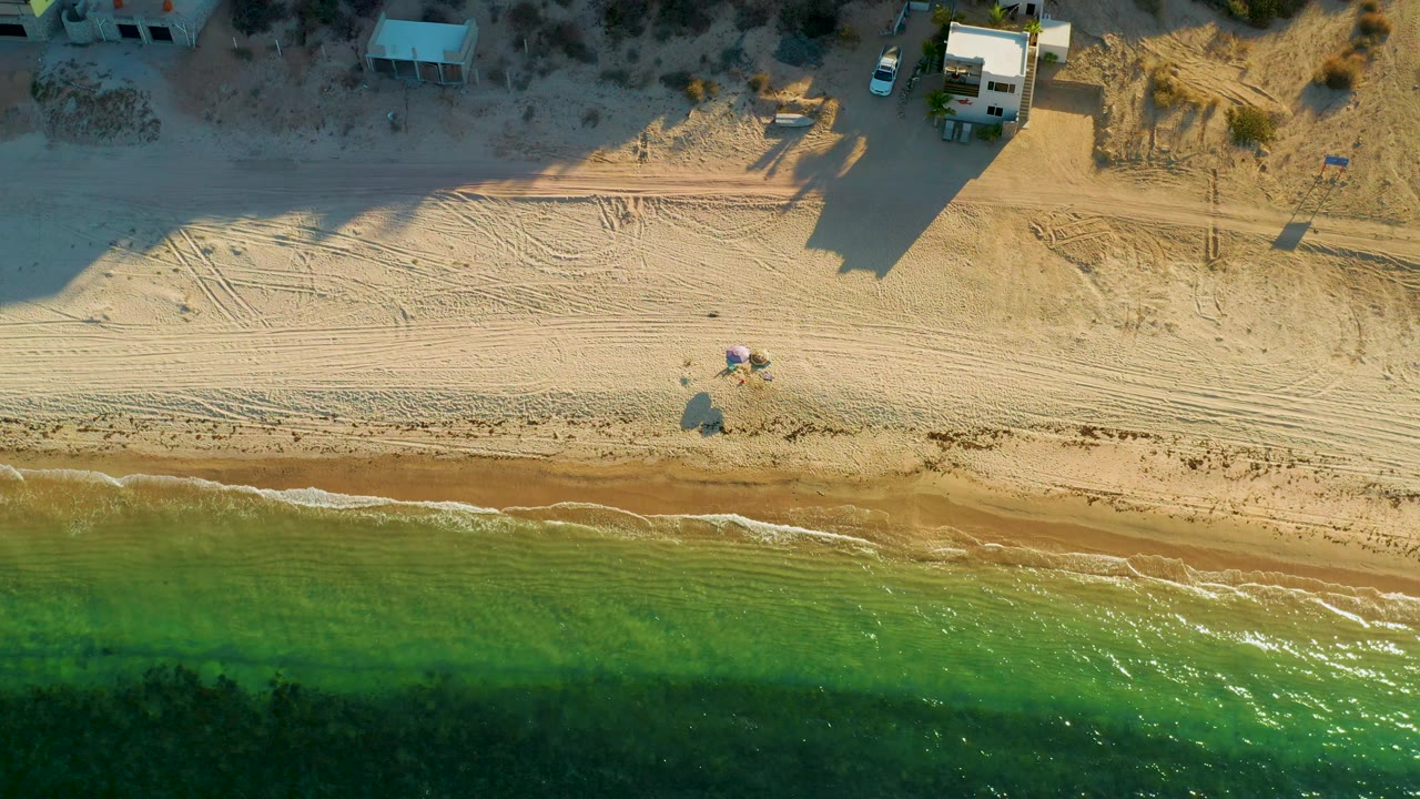 Bird's-eye frame over the El Sargento La Ventana coast, with clear green water along the lower edge, empty sand through the middle, houses at the upper edge, and no visible people.