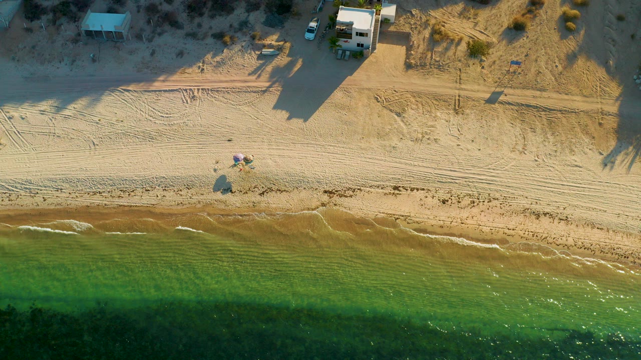Overhead frame of El Sargento La Ventana beachfront showing green water below, a thin surf edge, pale sand with tire tracks, small structures at the top, and no visible people.