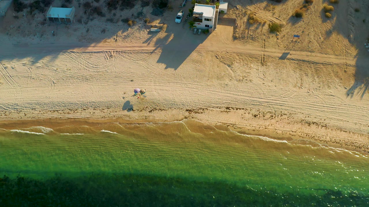 Aerial frame over El Sargento La Ventana with green water along the lower edge, a tan shoreline, tracked sand, small beach gear near center, and no visible people.