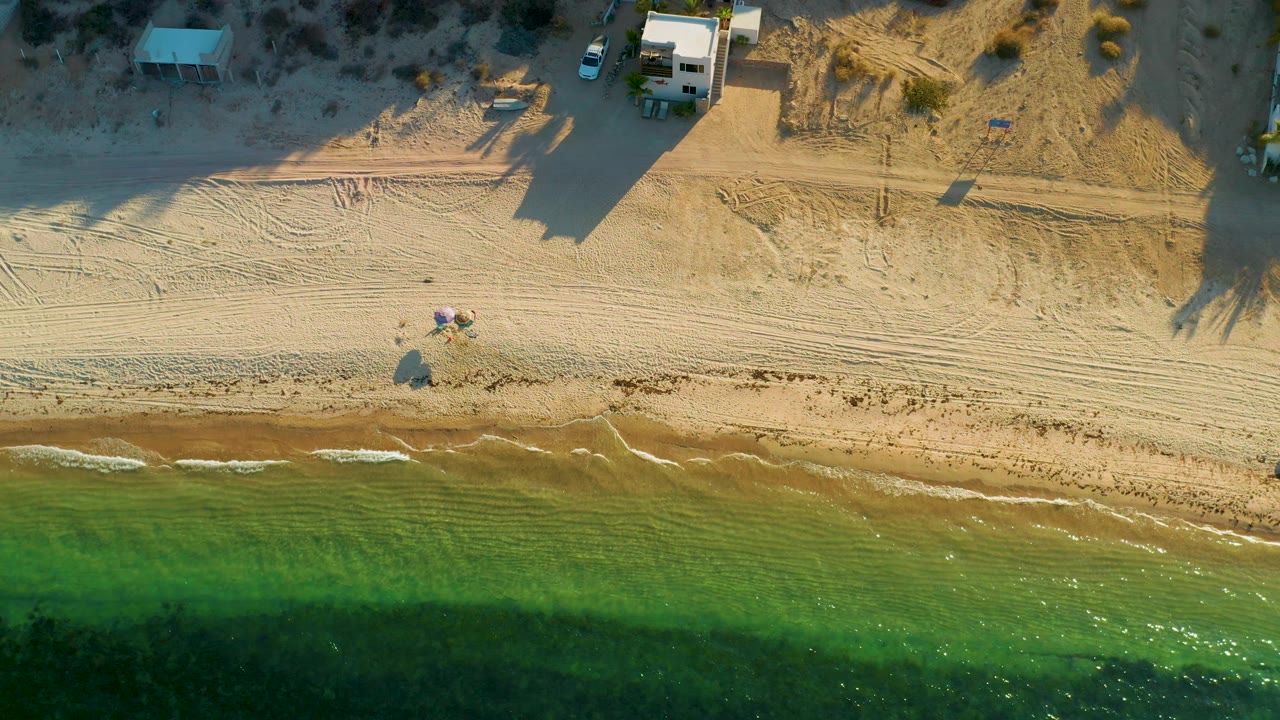 Top-down view of the El Sargento La Ventana shoreline with the surf band slightly lower in frame, tracked sand across the middle, small structures above, and no visible people.