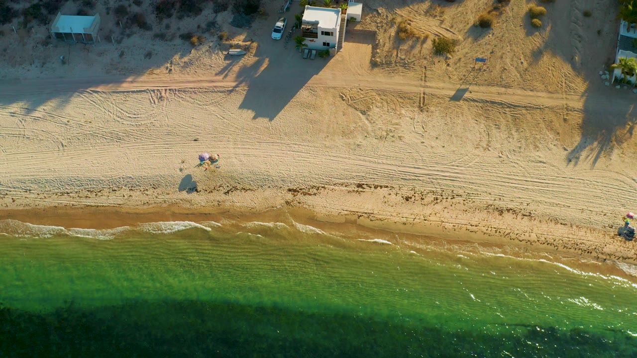 Drone view of sandy beachfront with green water, beach tracks, small gear, and building shadows.