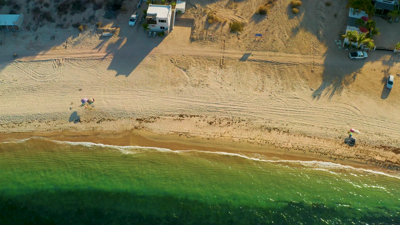 Aerial view in El Sargento La Ventana, Mexico showing shallow green water, a thin wave edge, raked or tire-marked sand, small shoreline objects, vehicles, buildings, and no visible people.