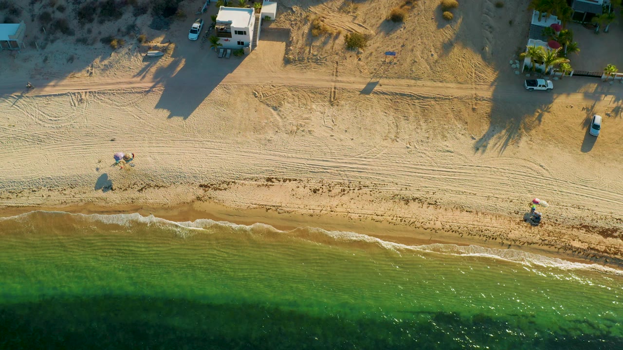 Top-down shoreline frame from El Sargento La Ventana, Mexico with the surf line crossing the lower third, tracked sand in the middle, beachside structures and vehicles above, and no visible people.