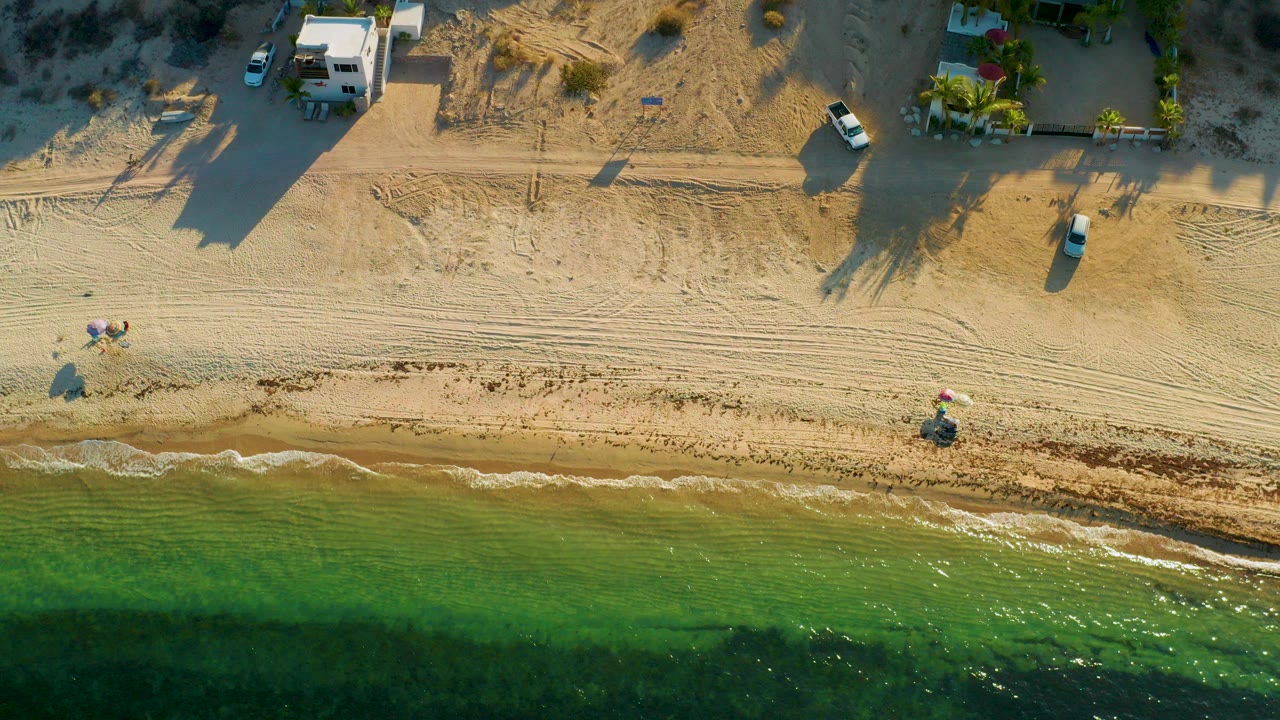 Aerial frame over El Sargento La Ventana, Mexico, showing green water at the bottom, a sandy beach with tire tracks, parked vehicles, and low buildings casting long shadows.
