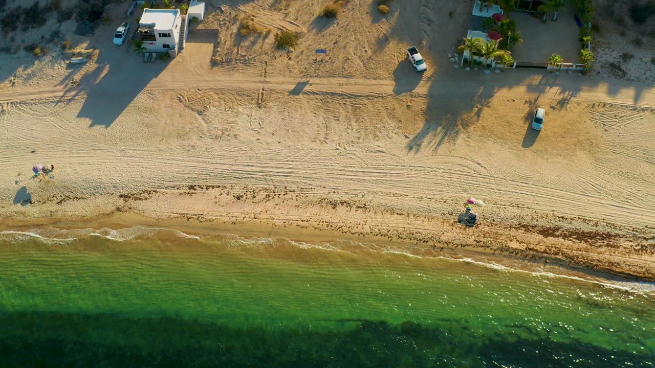Aerial frame over El Sargento La Ventana, Mexico, showing a narrow wave line on the beach, darker green water below, and several parked vehicles near beachside buildings above.