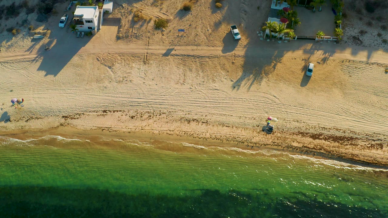 Top-down beach scene with small surf, sand tracks, vehicles, and coastal buildings.