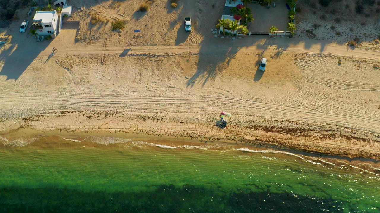 Overhead view in El Sargento La Ventana, Mexico, showing green shallow water below a tan sand beach with tire tracks, white shoreline buildings, parked vehicles, and no visible people.