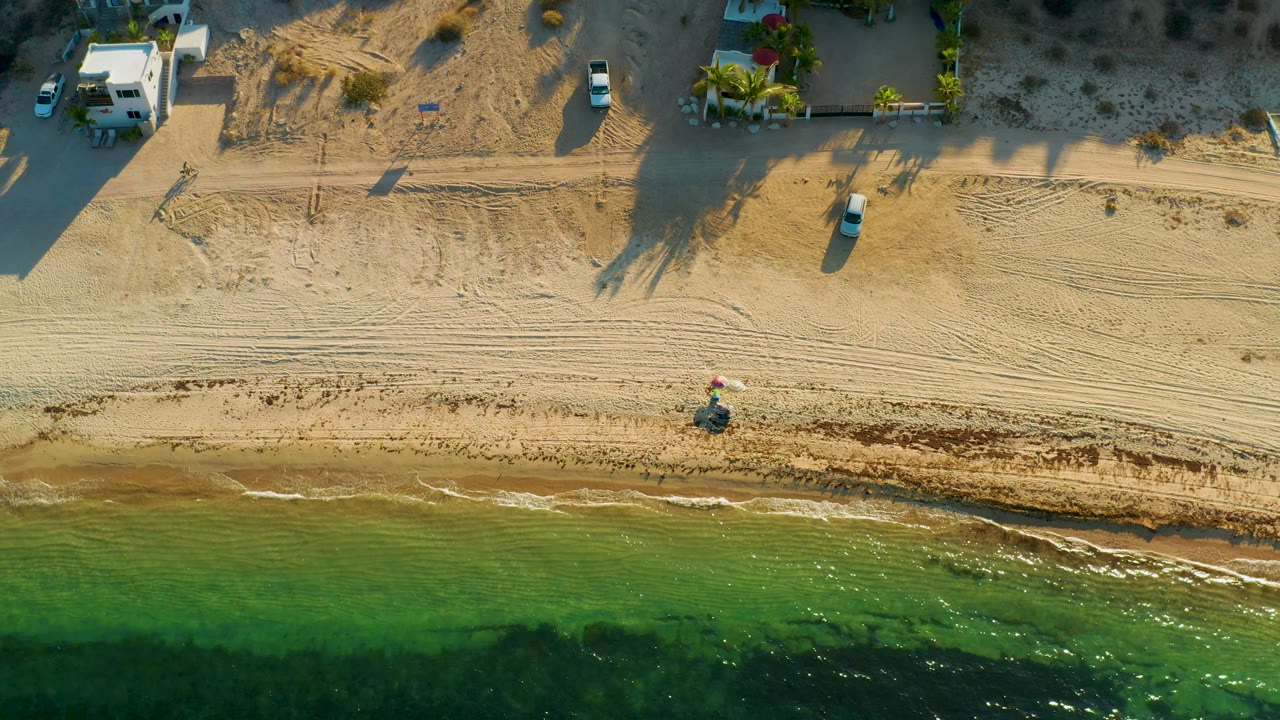 Top-down El Sargento La Ventana beach view with green sea in the lower third, raked sand and tire tracks through the center, buildings and parked vehicles above, and no visible people.