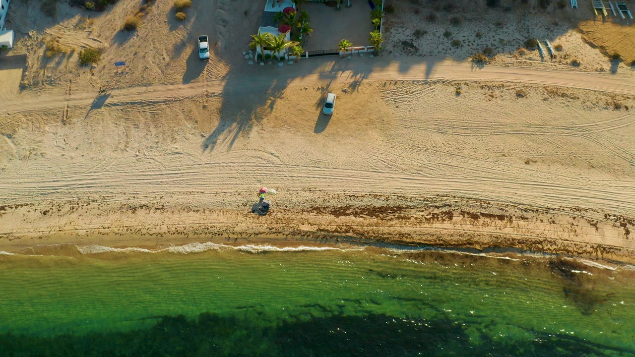 Overhead frame in El Sargento La Ventana, Mexico showing a small bicycle rider near the wet sand, green shallow water, beach tire tracks, parked vehicles, and beach-edge homes.