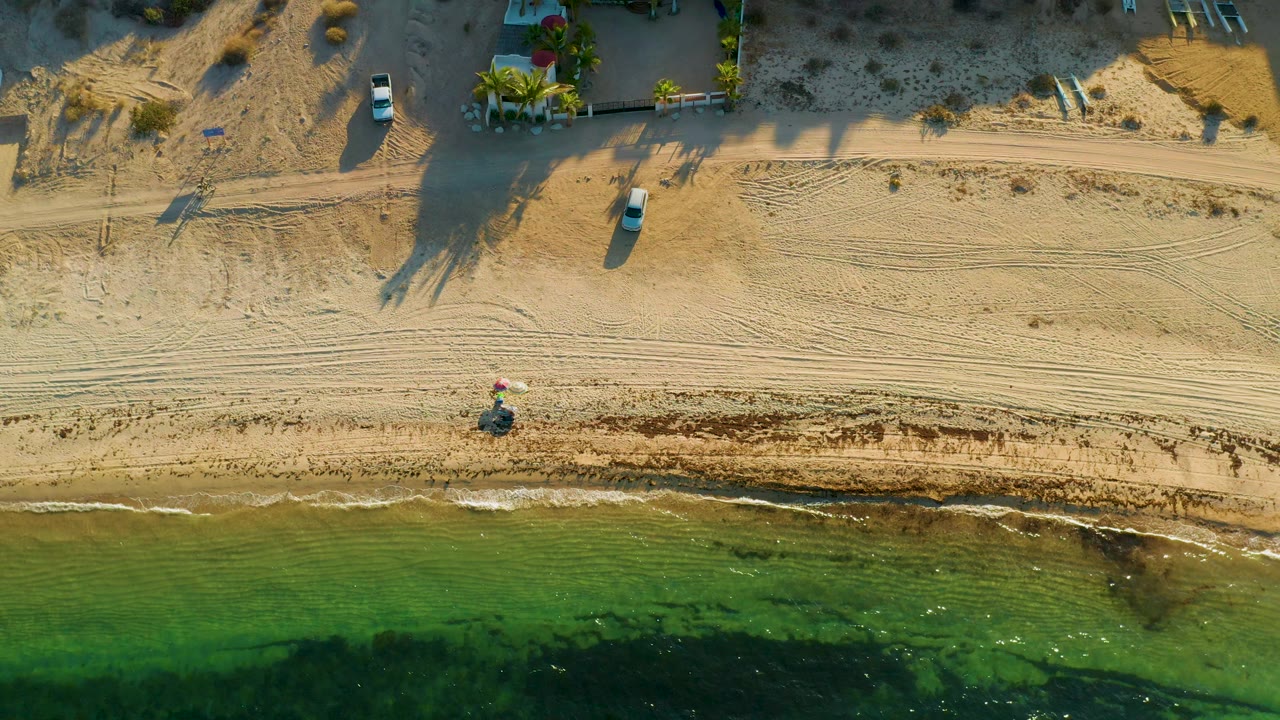 Top-down frame in El Sargento La Ventana, Mexico showing the bicycle rider near the lower sand band, with darker seaweed patches in the green water and beach homes partly visible above.