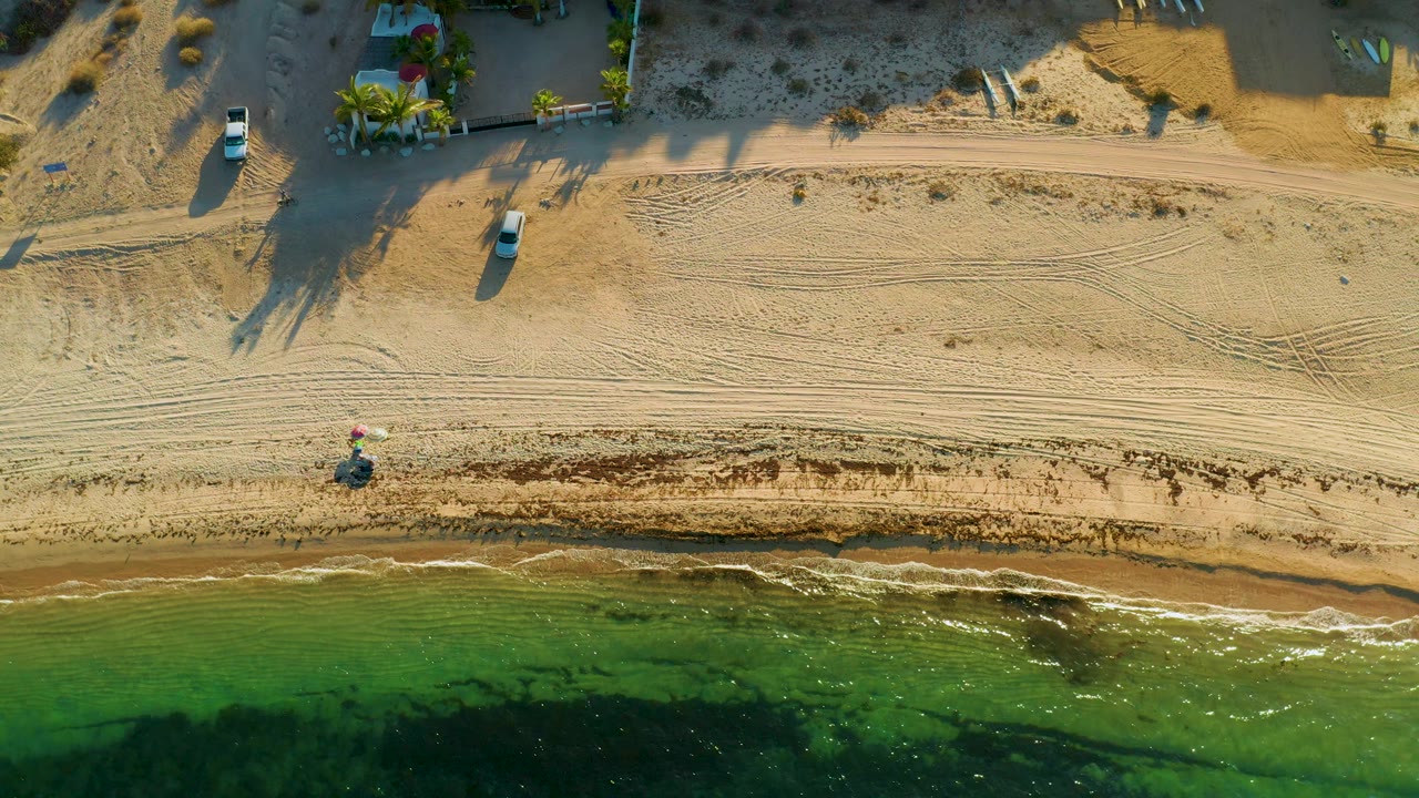 Aerial frame over El Sargento La Ventana, Mexico showing shallow green water, a narrow wet shoreline, patterned tire tracks, a sandy road, vehicles, and no visible people.