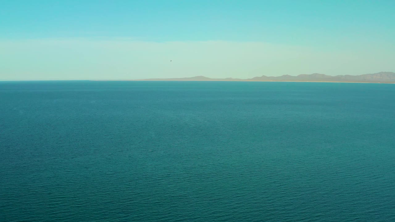 Calm blue sea stretches toward hazy tan mountains under a clear pale sky in El Sargento La Ventana, Mexico, with no visible people.