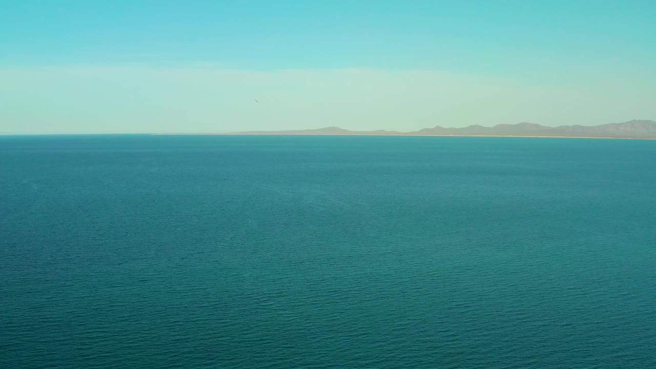 A large blue body of water leads to a distant shoreline and muted mountain shapes in El Sargento La Ventana, Mexico, with no visible people.