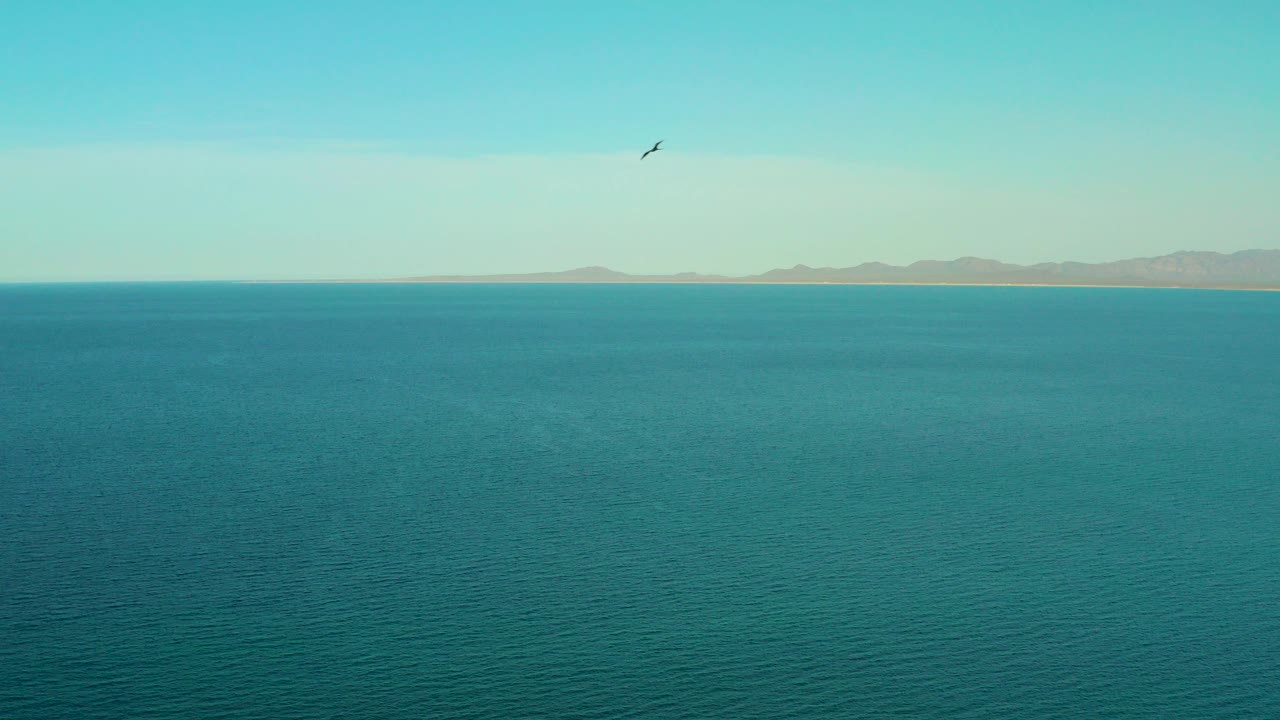 A wide view over blue water near El Sargento La Ventana, Mexico, with distant low mountains on the horizon, a small bird high in the sky, and no visible people.