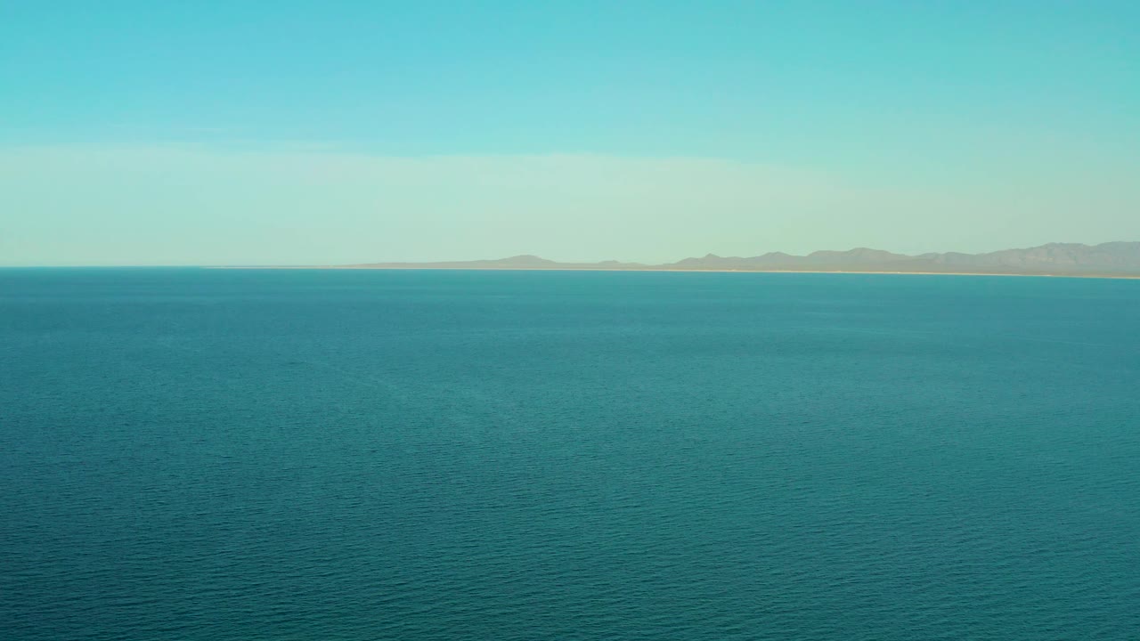 At 0:02, teal-blue ocean fills most of the frame while distant low mountains sit beneath the pale sky in El Sargento La Ventana, Mexico, with no visible people.