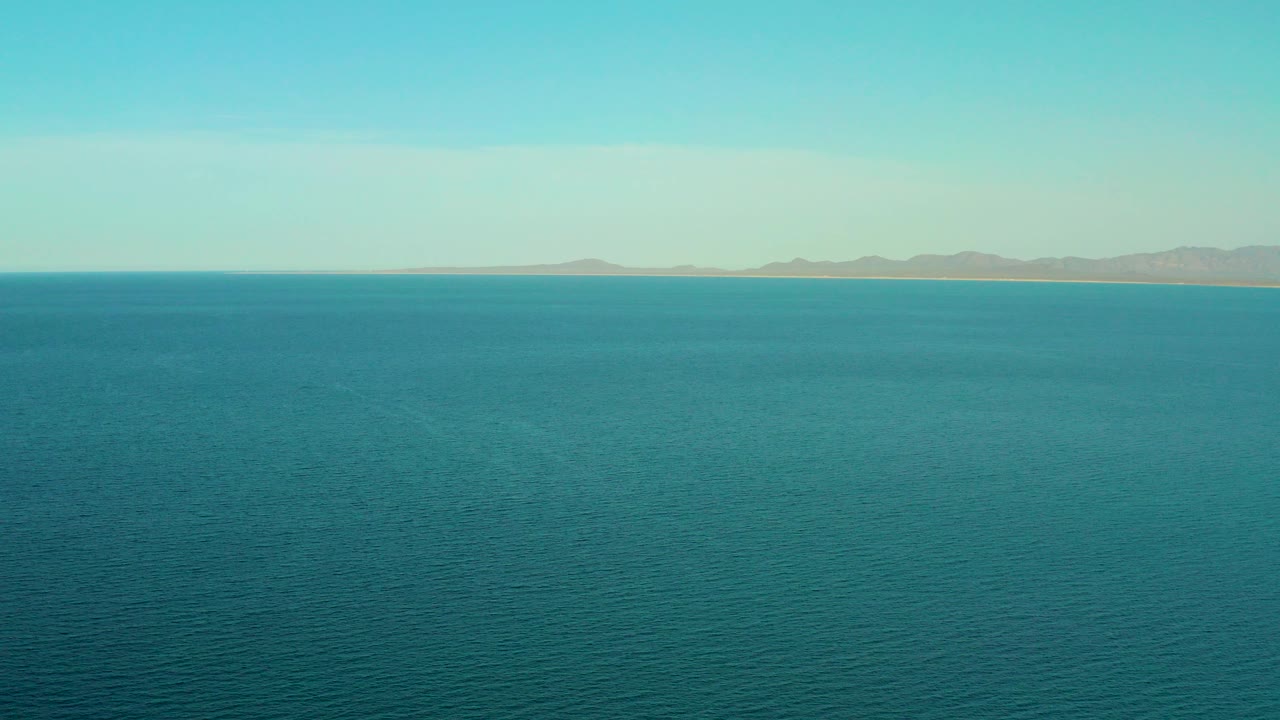 Opening frame of blue-green ocean near El Sargento La Ventana, Mexico, with a thin tan coastline and soft mountain shapes on the horizon, no visible people.