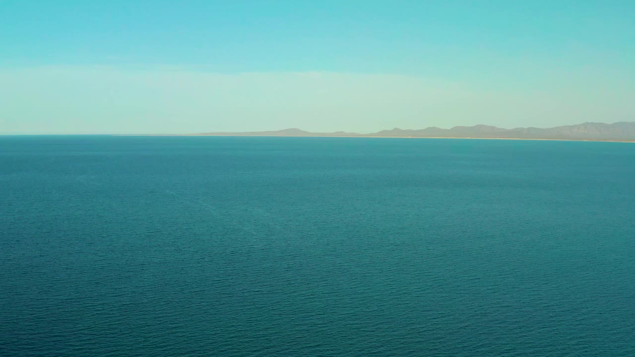Early frame over open ocean at El Sargento La Ventana, Mexico, with subtle water texture in the foreground and hazy mountains set far back, no visible people.