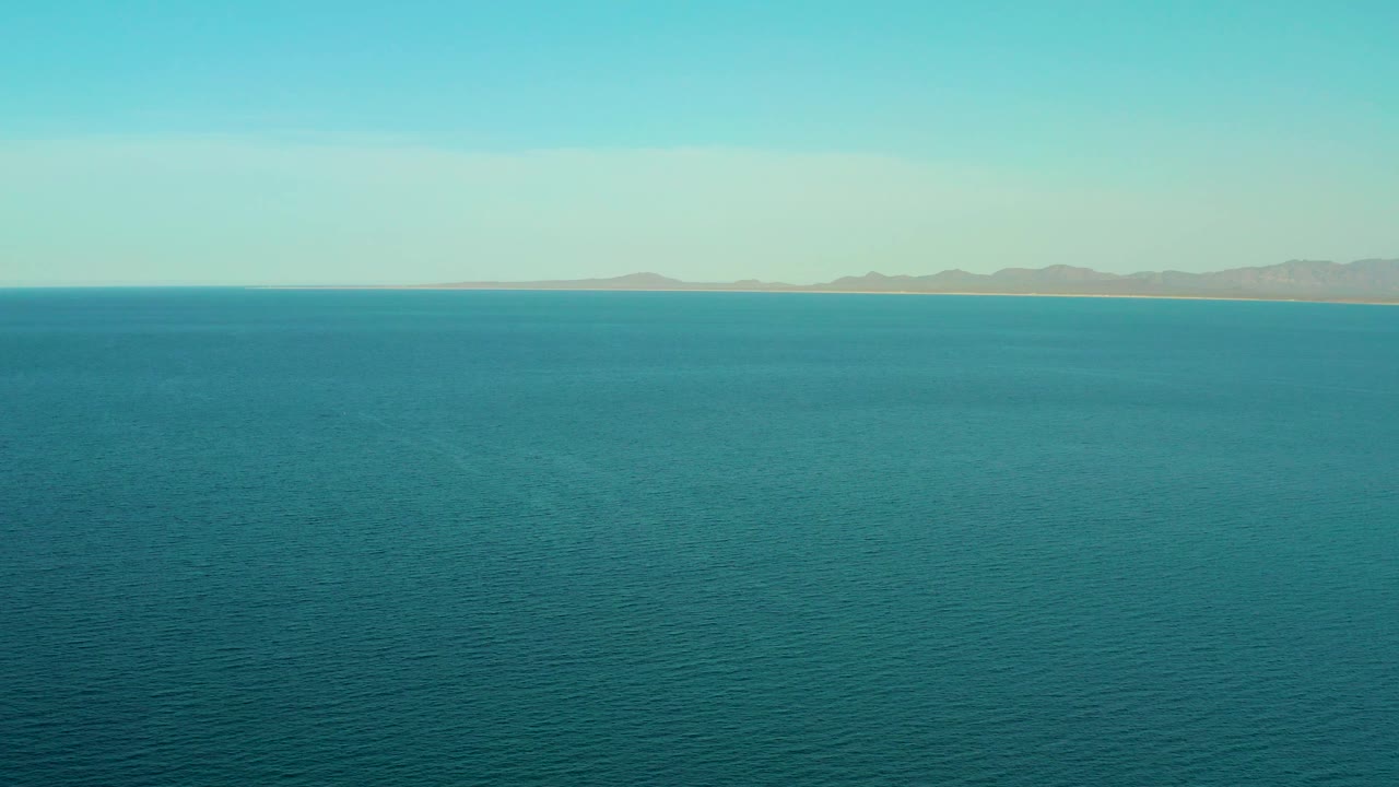 Mid-clip frame from above the ocean at El Sargento La Ventana, Mexico, showing deeper teal water below and a tan coastal band beneath muted mountains, no visible people.